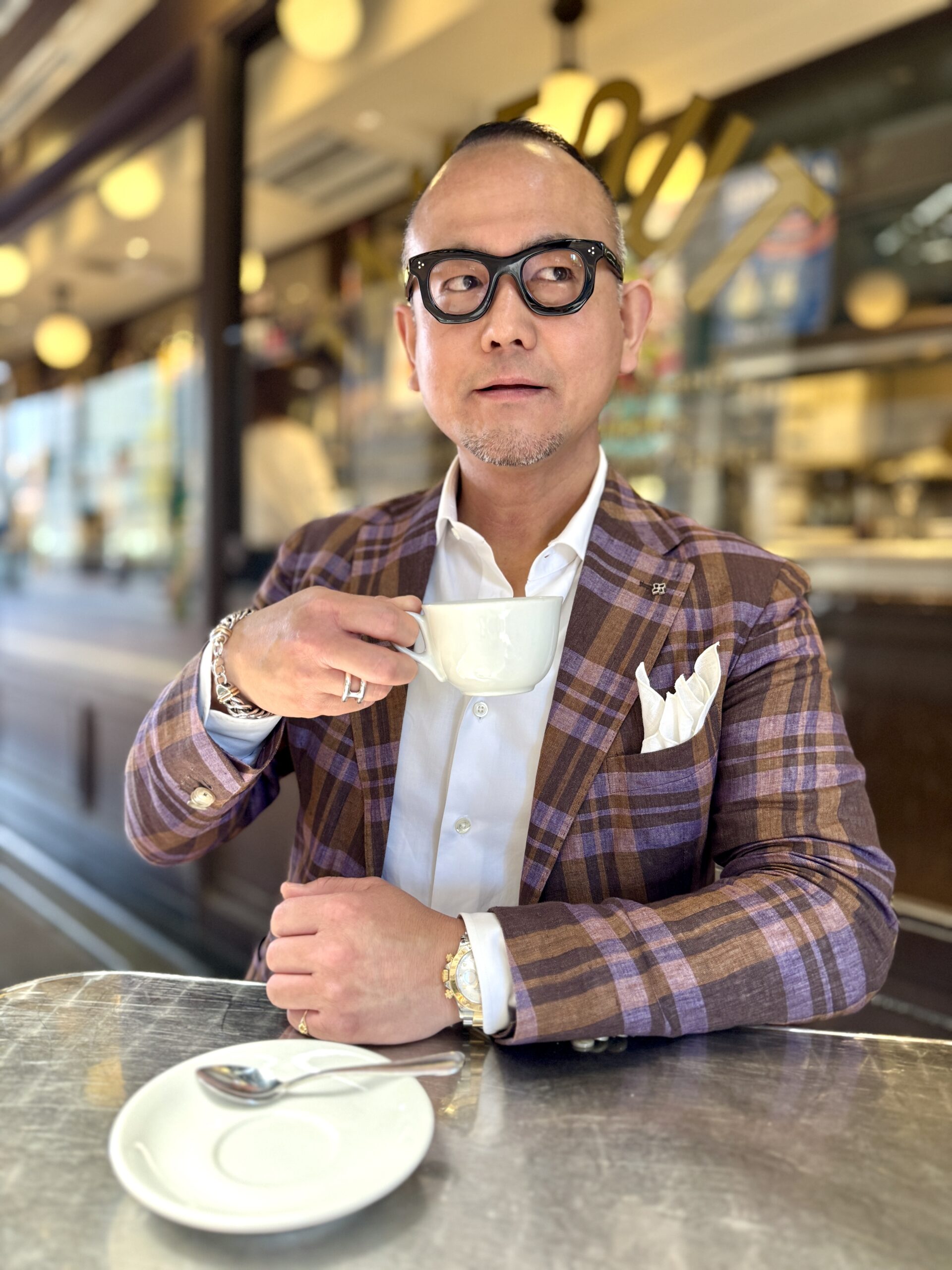 チェック柄のジャケット姿でカフェに座るコバヤシシゲユキ